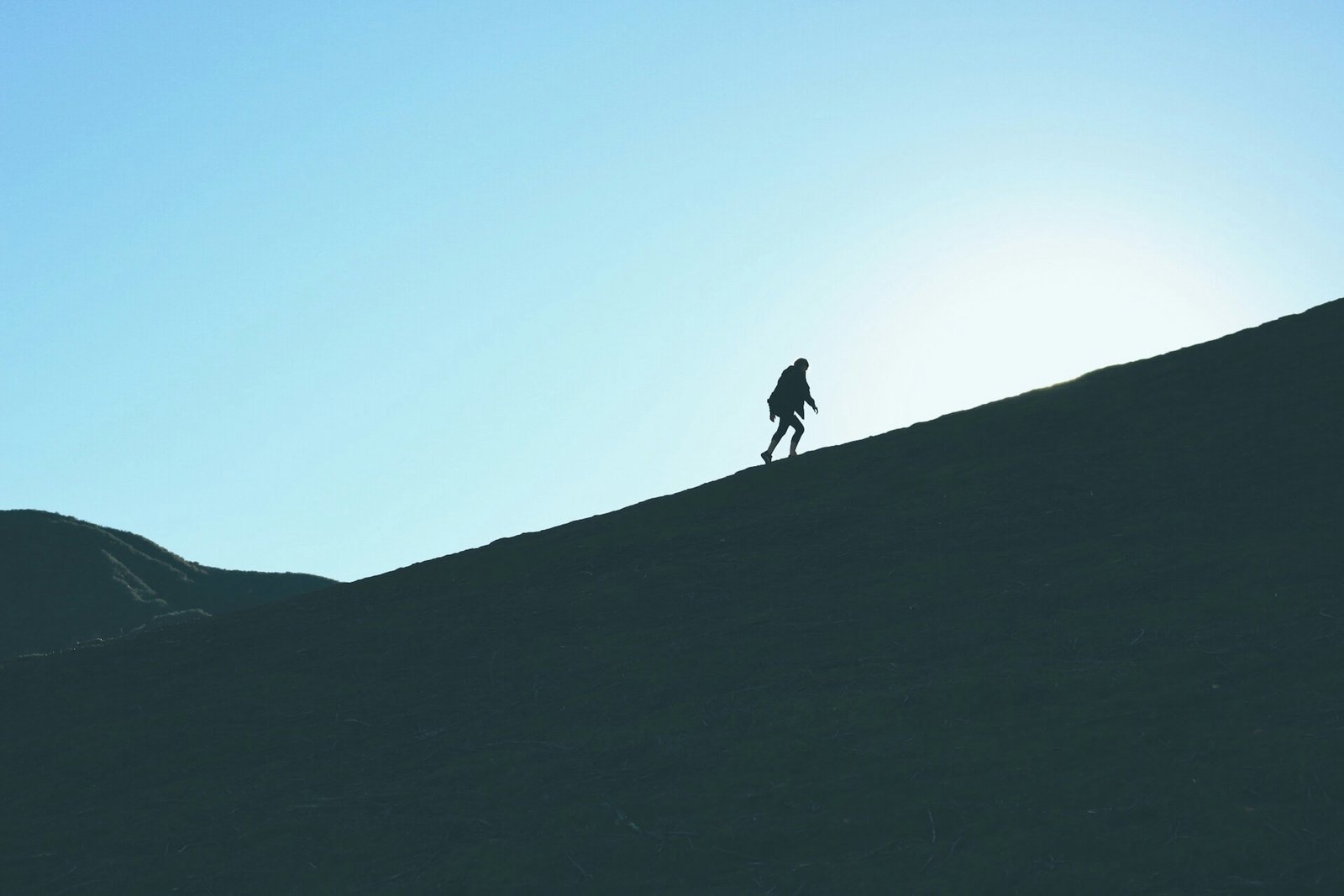 A lone figure walking steadily up a steep hillside against a clear sky